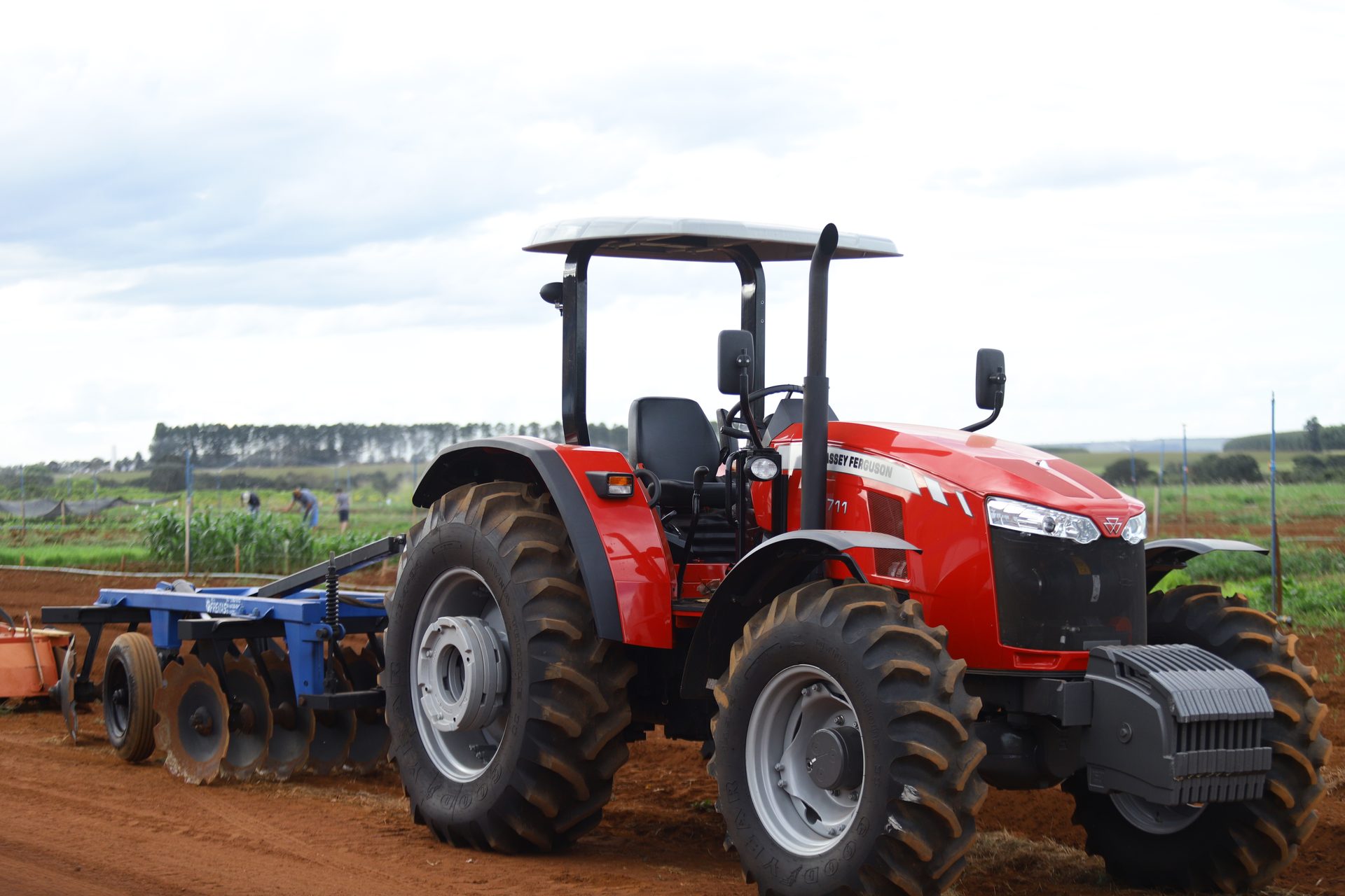 Trator agrícola vermelho de grande porte, sem cabine fechada (apenas cobertura protetora), acoplado a um implemento agrícola de grade niveladora azul, estacionado sobre estrada de terra vermelha.