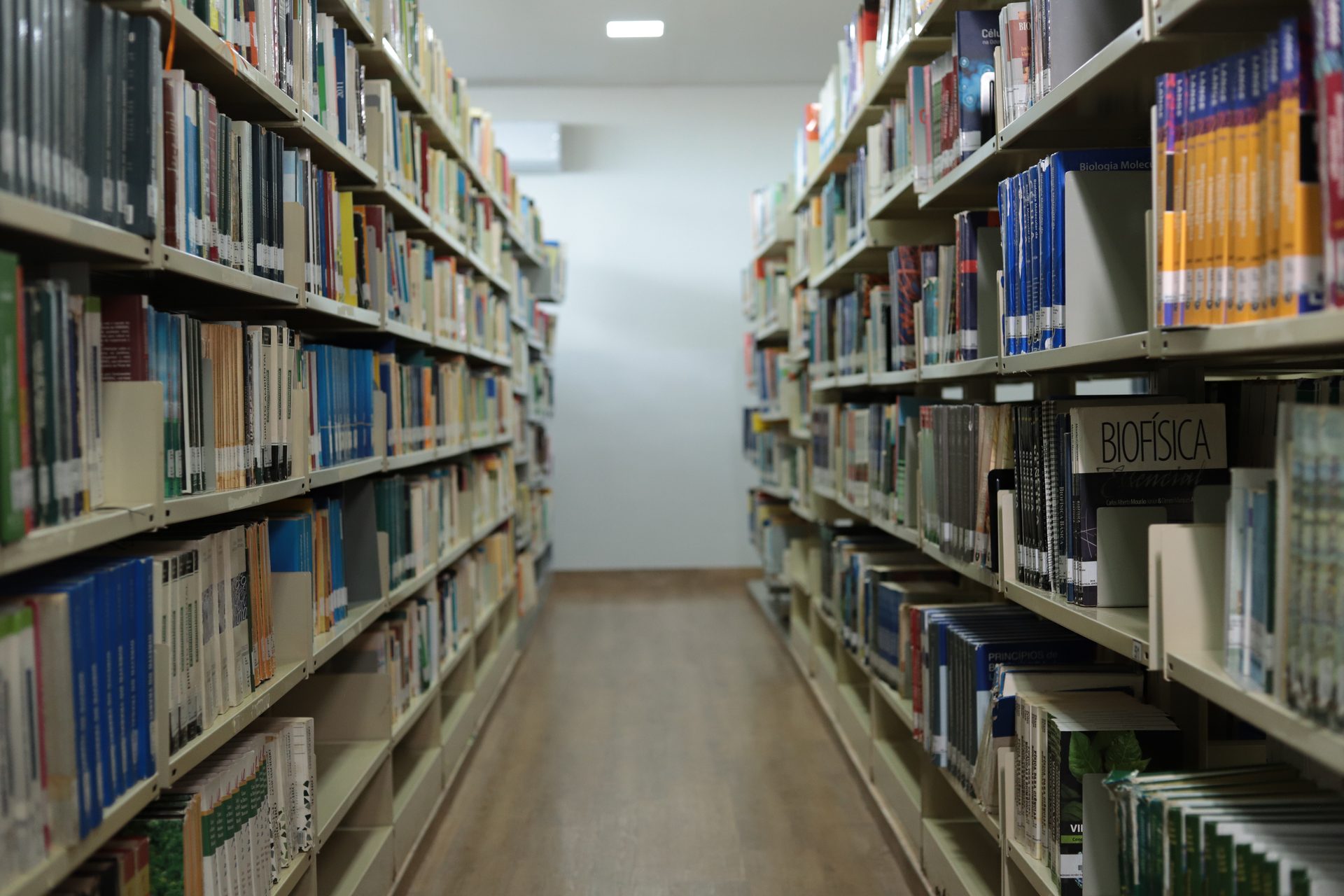 Corredor entre duas estantes altas de biblioteca, repletas de livros organizados por lombada, com títulos como 'Biofísica' visíveis nas laterais dos exemplares.