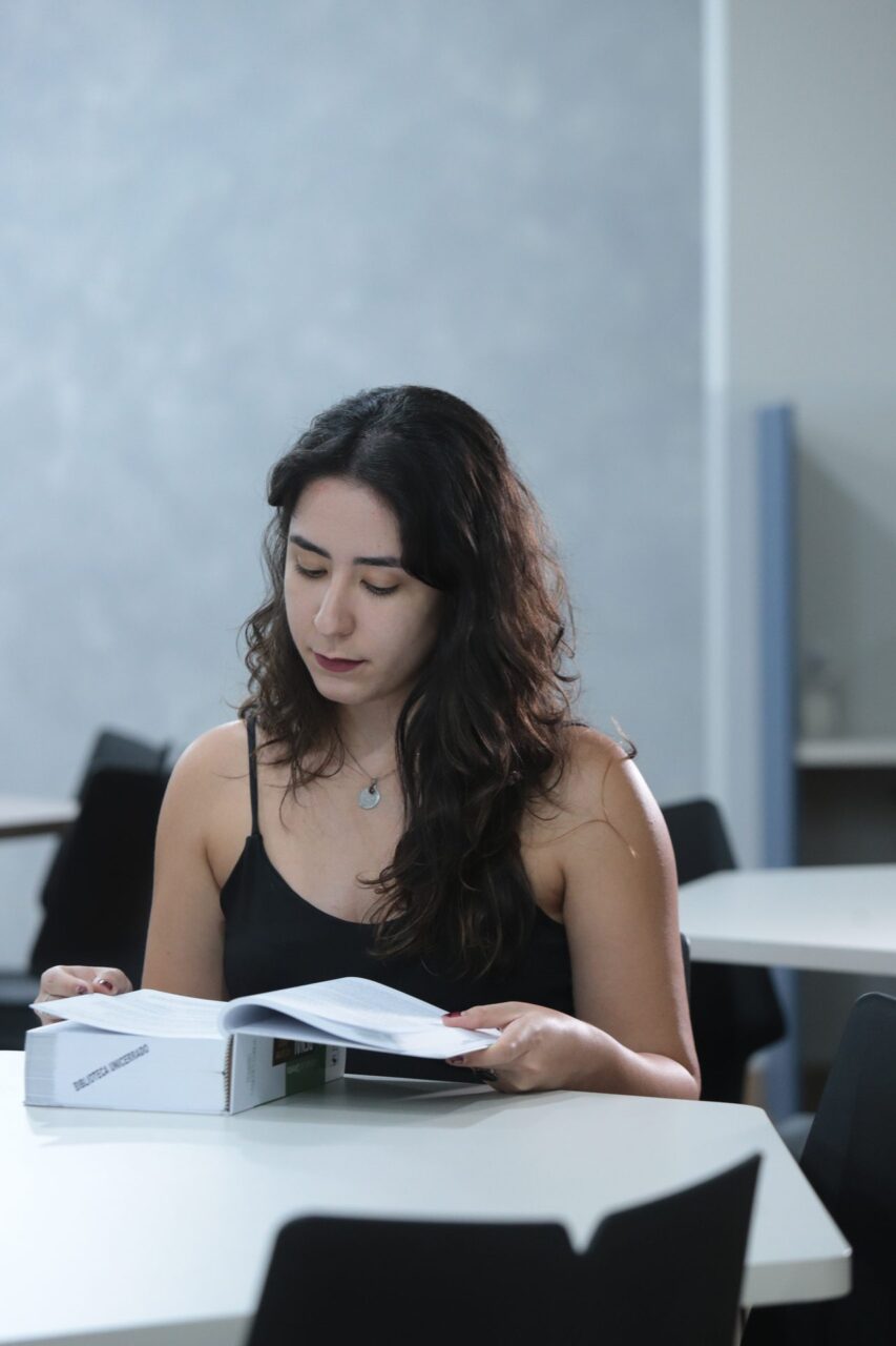 Mulher de cabelos longos e castanhos, sentada a uma mesa branca, folheando um livro grosso com etiqueta 'Biblioteca Universitária' na lombada.