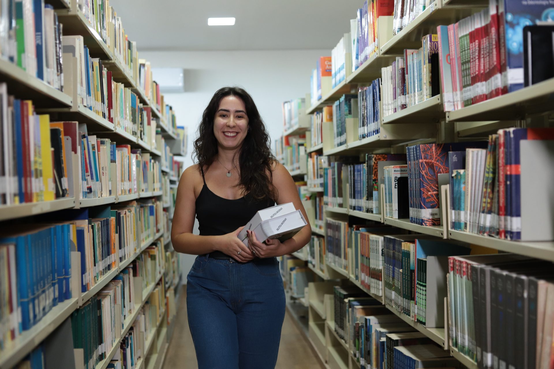 Mulher jovem sorridente, cabelos longos e ondulados, vestida com blusa preta e calça jeans, caminha em um corredor entre estantes altas de livros, carregando alguns livros nas mãos.