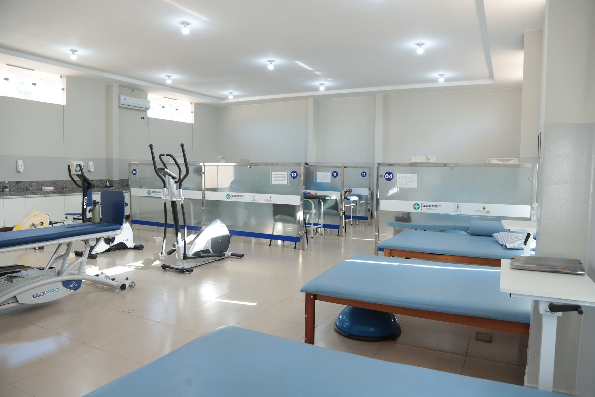 Ambiente amplo de clínica com várias macas de atendimento com estofado azul e estrutura de madeira, boxes individuais delimitados por divisórias de vidro/acrílico numeradas (10, 11, 12, 04), equipamentos de fisioterapia como elíptico e bicicleta ergométrica, bosu azul no chão, bancada com pia ao fundo e ar-condicionado de parede.
