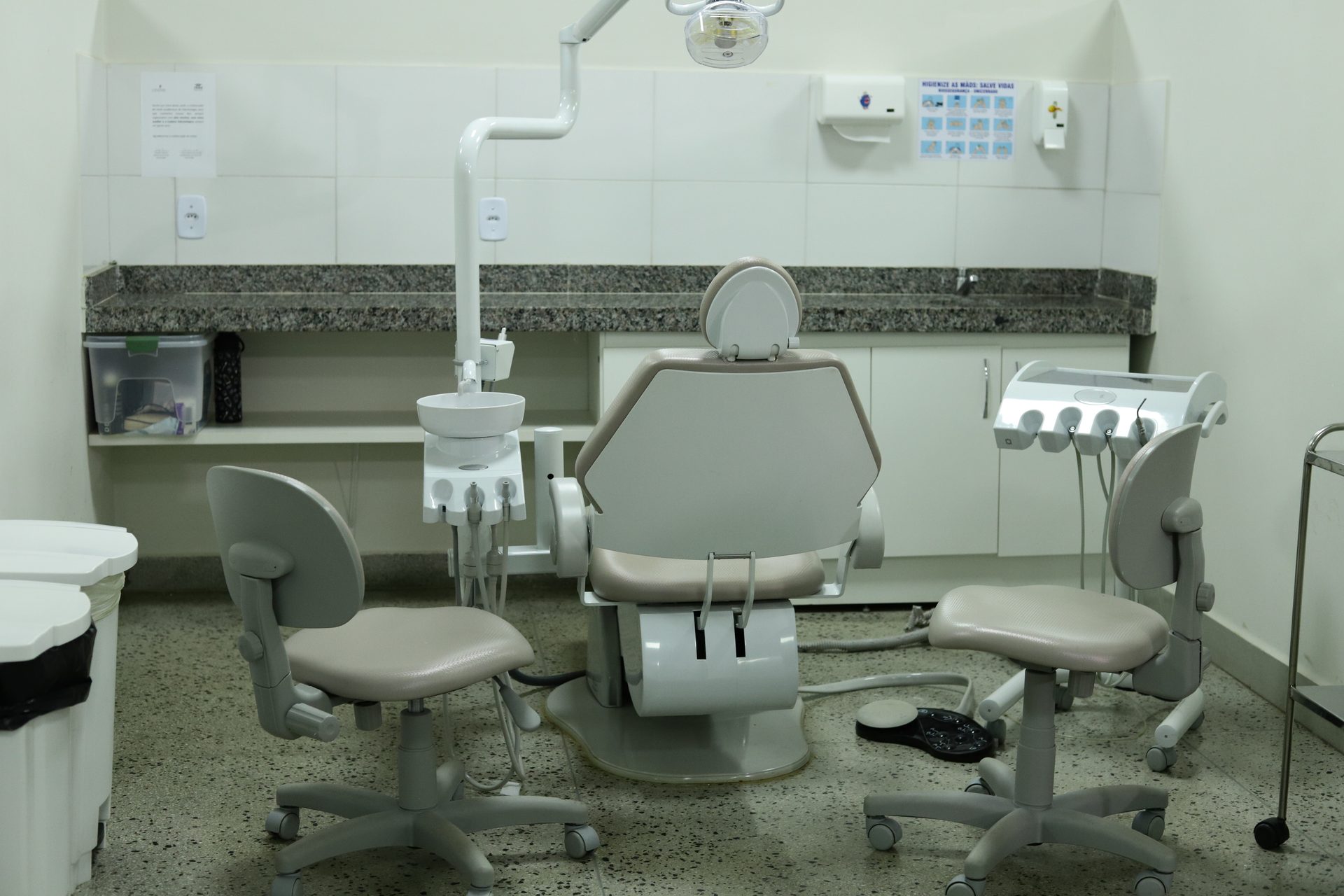 Consultório odontológico com cadeira reclinável central equipada com refletor, mocho e cadeira de apoio para dentista e auxiliar, bandeja com instrumentais suspensa à direita, pia com bancada de granito ao fundo, armários brancos embaixo da bancada, dispensadores de papel e álcool gel fixados na parede azulejada, cartazes informativos e um pedal de acionamento no chão.
