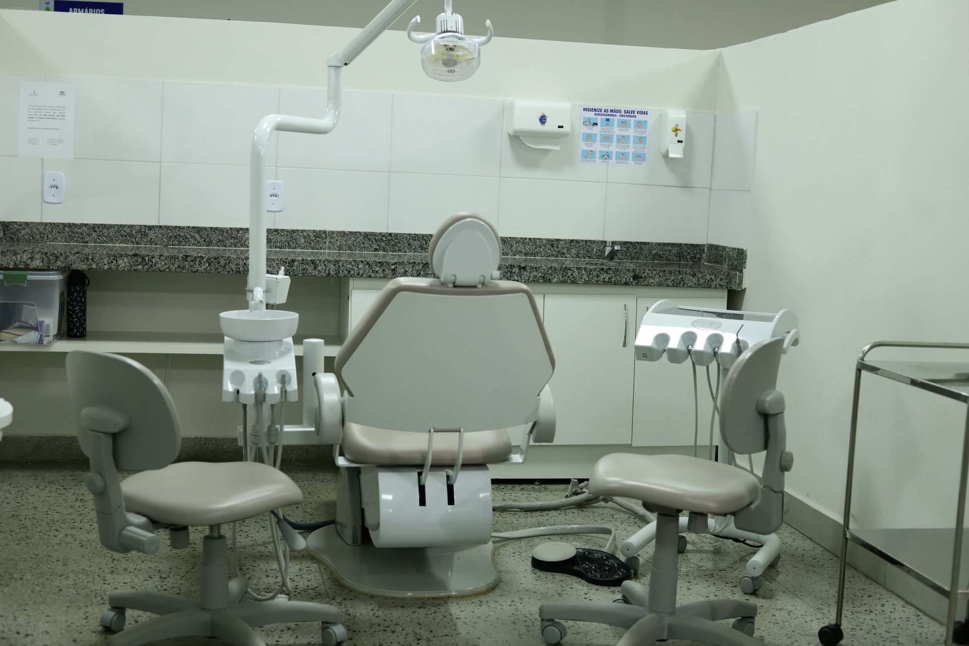 Consultório odontológico com cadeira de dentista central, equipada com refletor no teto, braço de sugador e seringa tríplice acoplados.