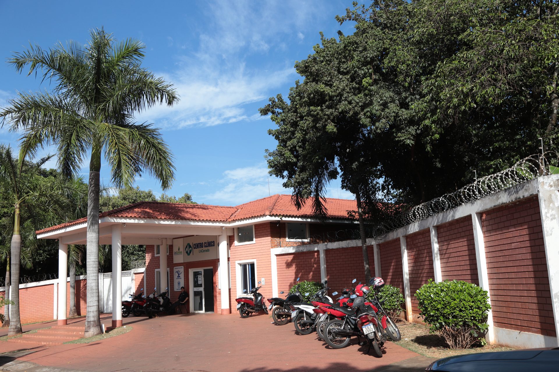 Fachada de um prédio térreo com telhado de telhas cerâmicas e paredes em tijolo aparente, com placa 'Centro Clínico' na entrada.