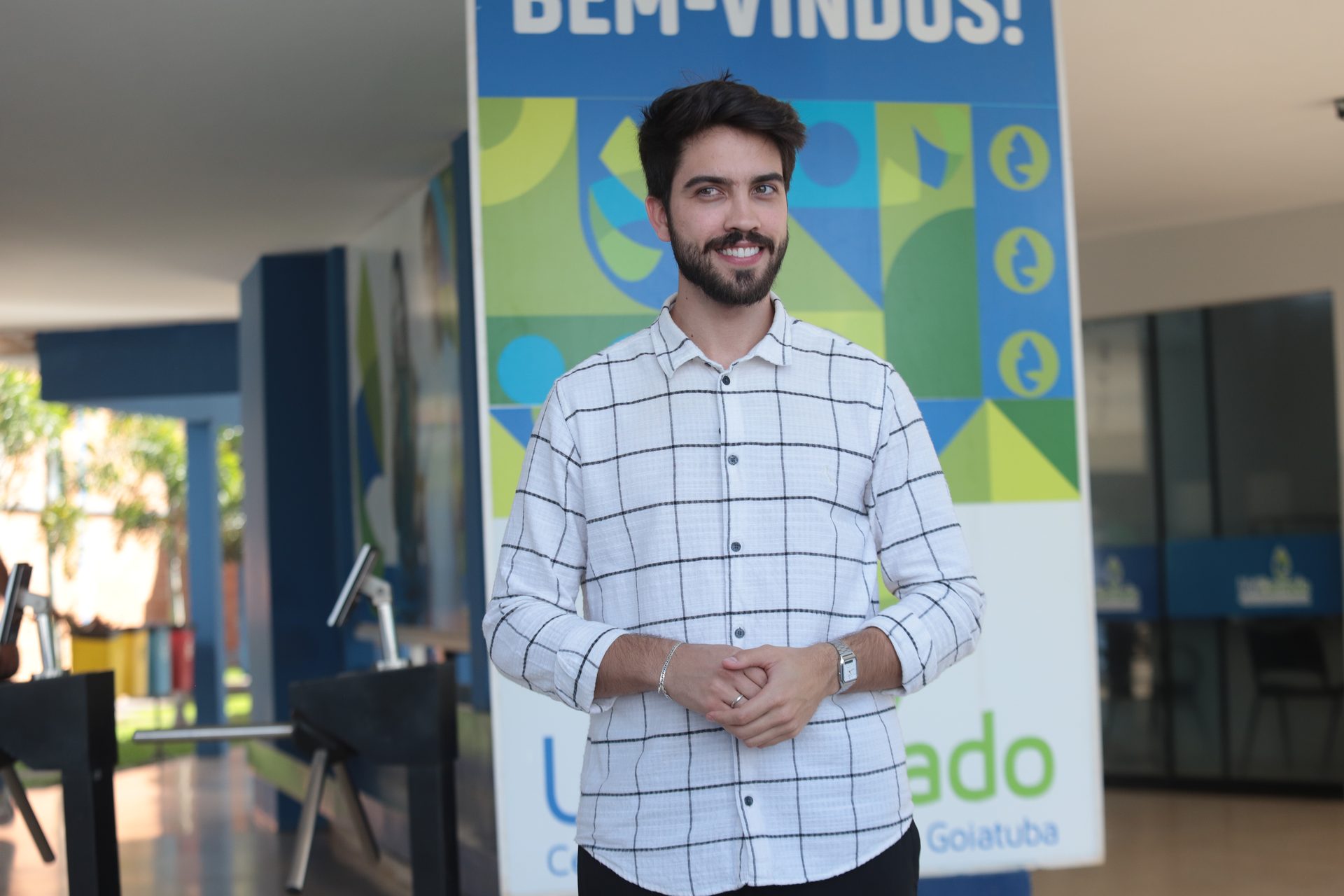 Homem de barba e cabelo curto, sorridente, posa em ambiente interno com as mãos entrelaçadas à frente do corpo.