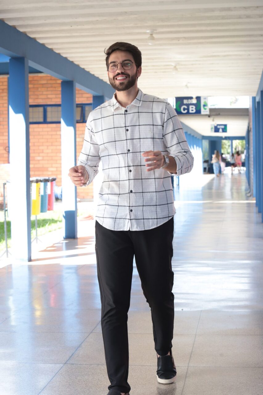 Homem jovem, sorridente, de óculos, barba e cabelo curto escuro, caminha por um corredor coberto de um prédio institucional.