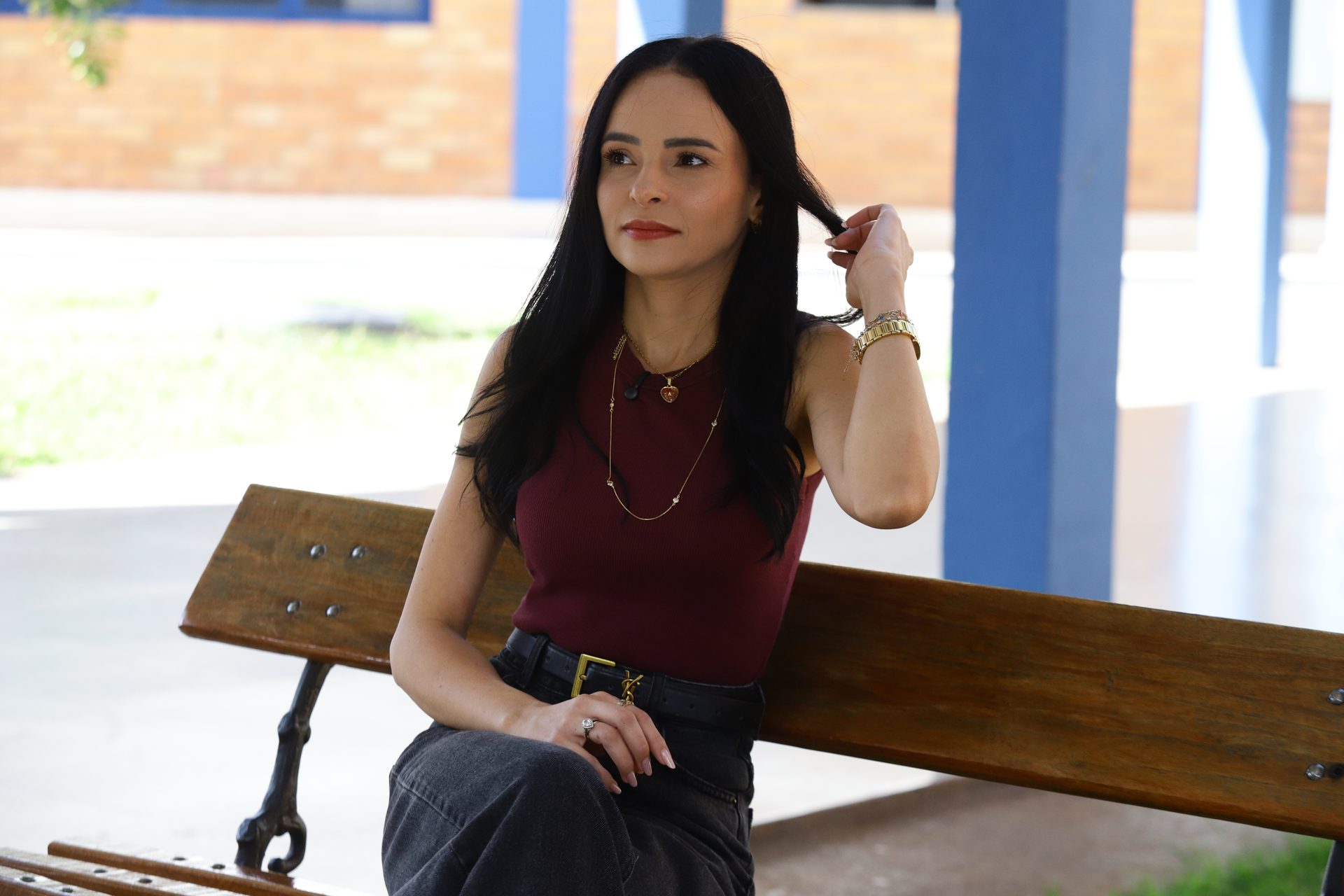 Mulher de cabelo longo e escuro sentada em um banco de madeira com estrutura de ferro, em uma área externa coberta com pilares azuis e parede de tijolos aparentes ao fundo.