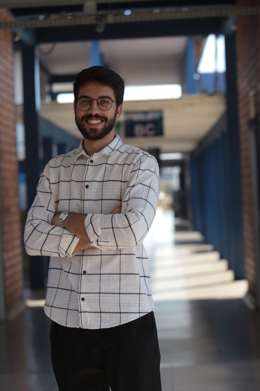 Retrato de um homem jovem, de cabelo e barba escuros, usando óculos de armação redonda, sorrindo com os braços cruzados.