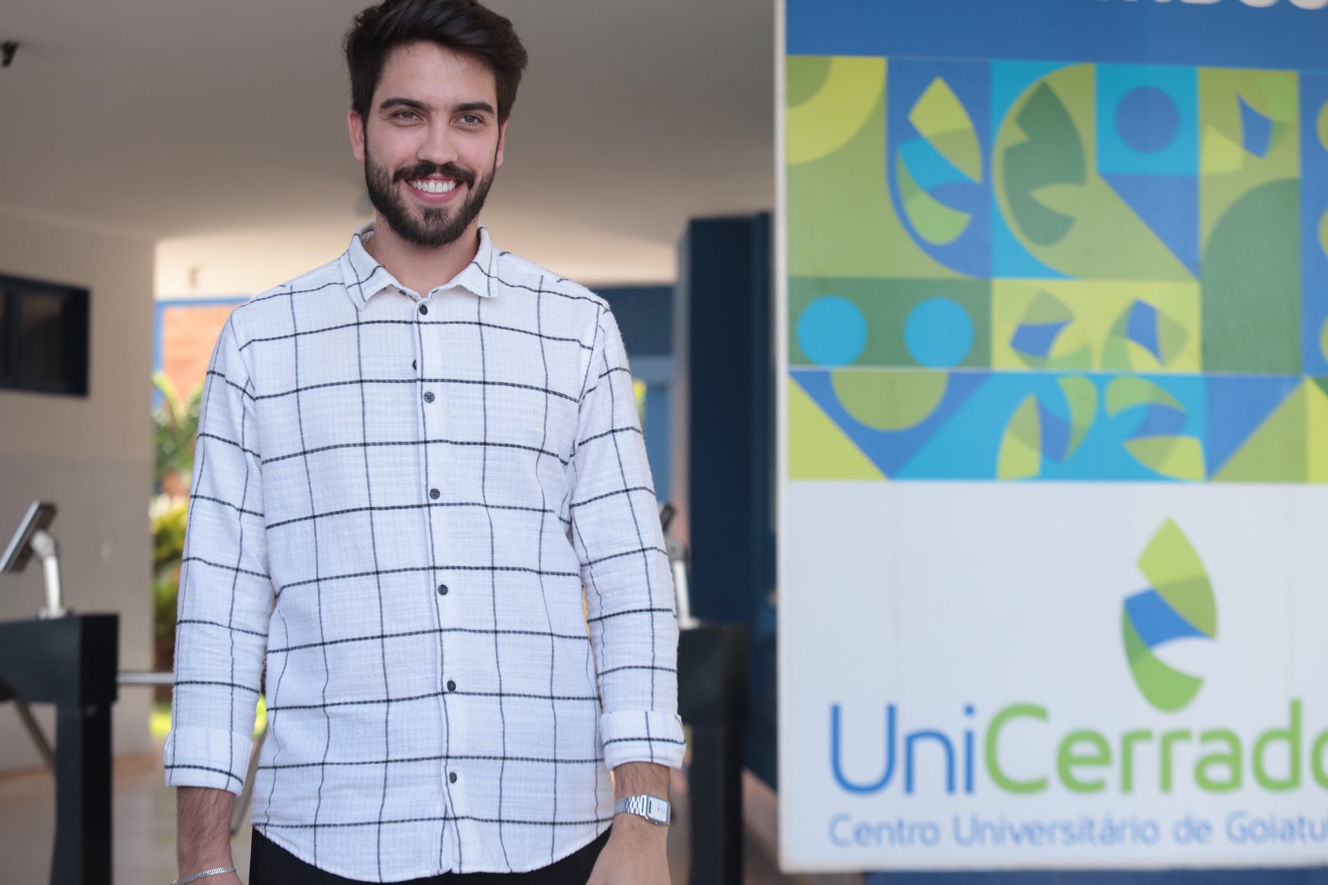 Retrato de um homem jovem, sorridente, com barba e cabelo curto escuro, vestindo camisa xadrez branca e preta de mangas compridas.