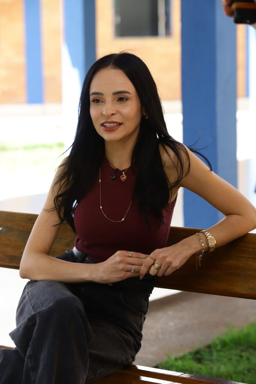 Retrato de uma mulher jovem sentada em um banco de madeira em área externa, com longos cabelos escuros soltos, vestindo blusa regata vinho, calça jeans escura e diversos acessórios (colar, anéis, relógio de pulso e pulseiras).