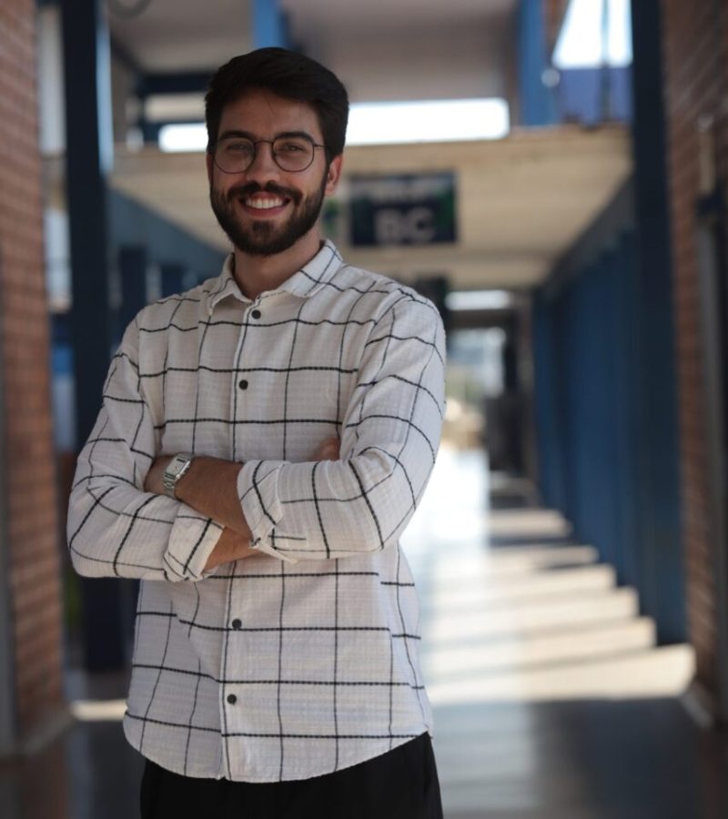 Retrato de um homem jovem, de cabelo e barba escuros, usando óculos de armação redonda, sorrindo com os braços cruzados.