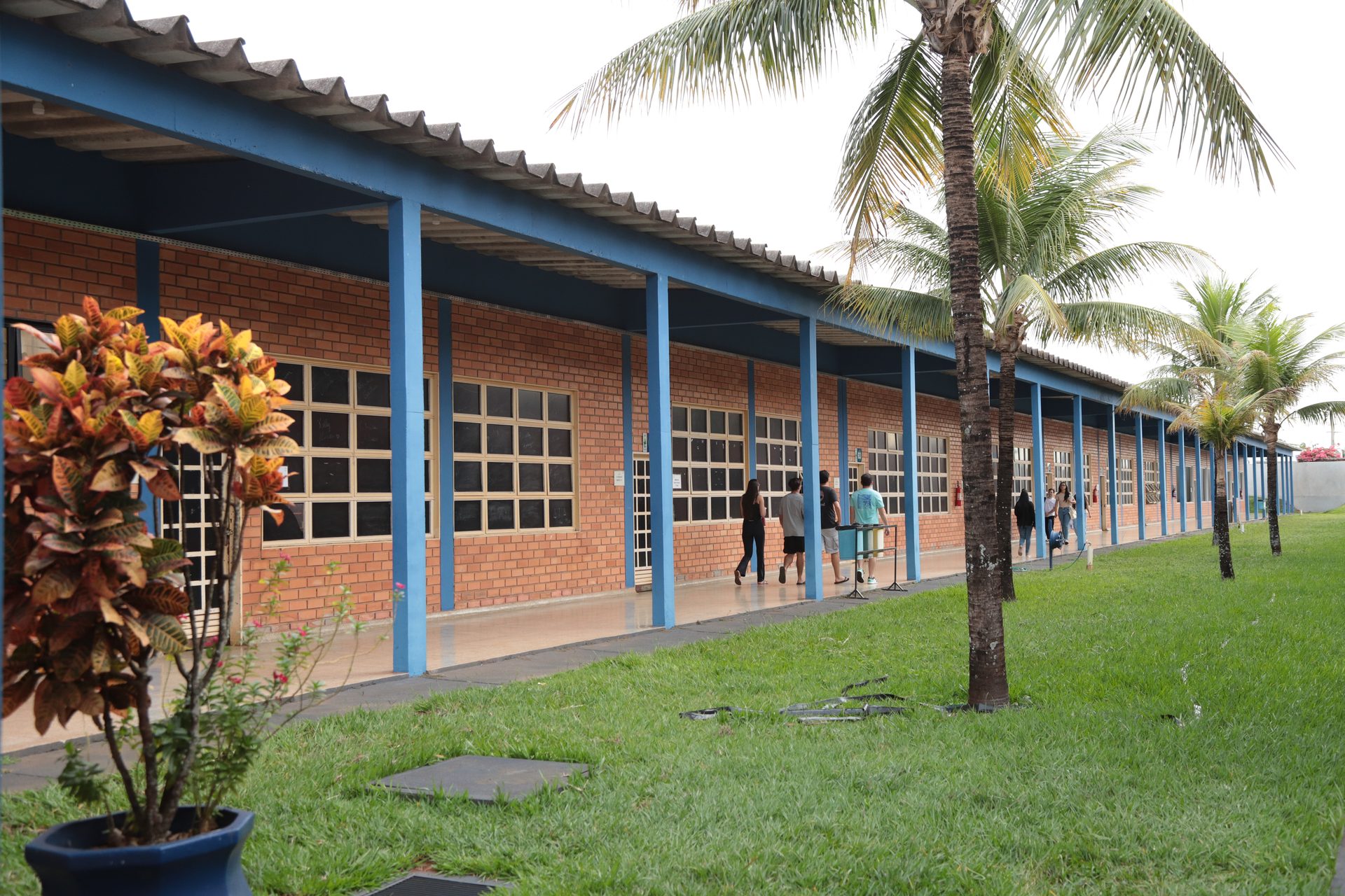 Fachada de um bloco de salas em tijolo aparente com estrutura e pilares azuis, cobertura de telhas onduladas e ampla varanda com piso claro.