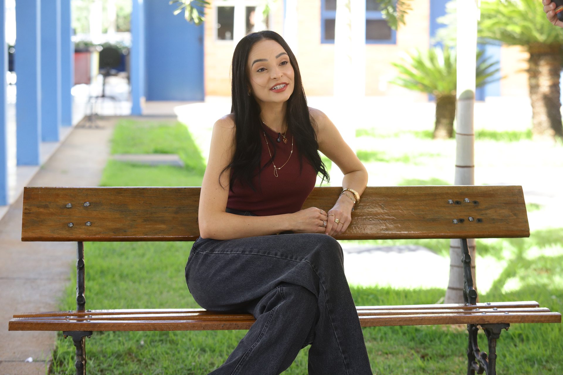 Mulher sentada em um banco de madeira e ferro em área externa do campus, vestindo blusa regata vinho e calça jeans, com os braços apoiados no encosto do banco e falando enquanto olha para o lado, em pose de entrevista ou depoimento.