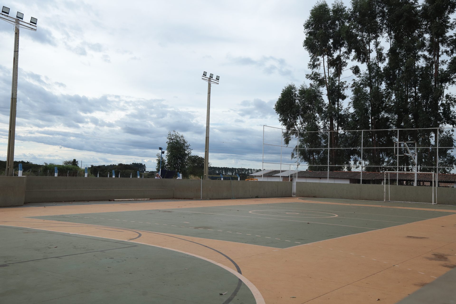 Quadra poliesportiva externa vazia, com piso pintado em verde e detalhes laranja/branco, cercada por alambrado e postes de iluminação.