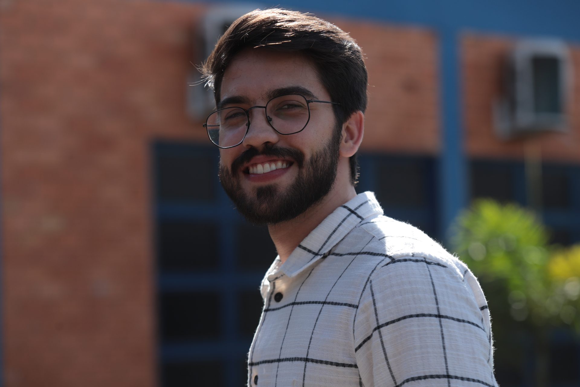 Retrato de um homem jovem, de cabelo curto e escuro, óculos de armação redonda e barba cheia, sorrindo e olhando para a câmera.