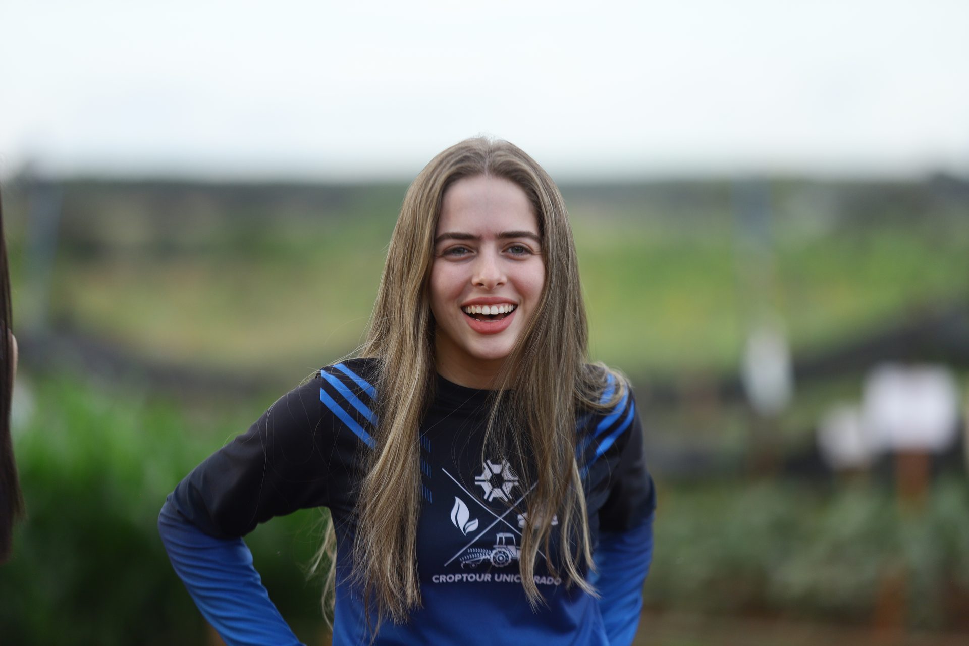 Retrato de uma jovem sorrindo, com cabelos longos e lisos, vestindo camiseta esportiva azul e preta com estampa 'CROPTOUR UNICERRADO' e um ícone de trator.
