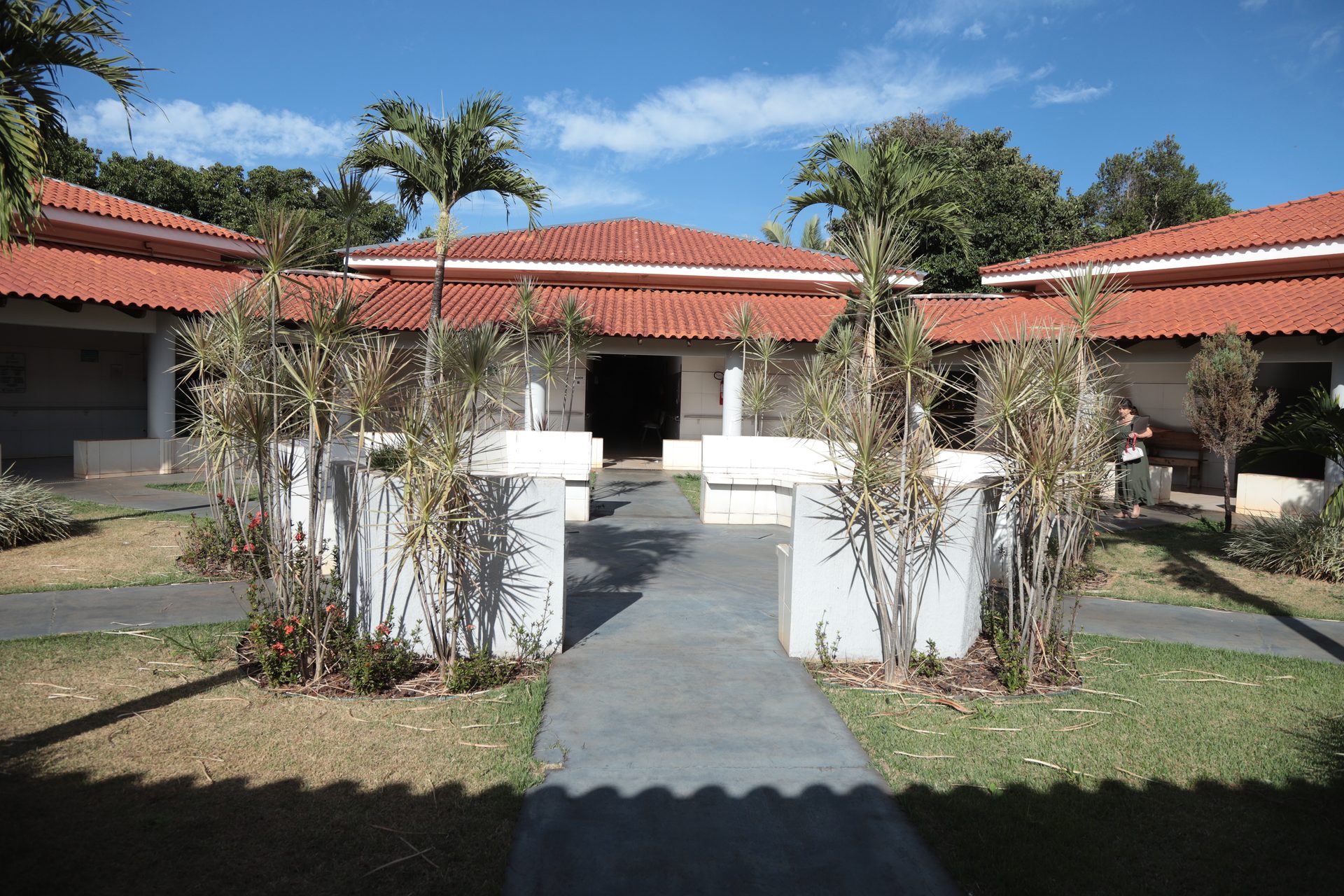 Vista externa de um conjunto de edificações térreas com telhado colonial em telhas cerâmicas, dispostas ao redor de um jardim central com palmeiras, plantas ornamentais de folhas pontiagudas e canteiros de grama.