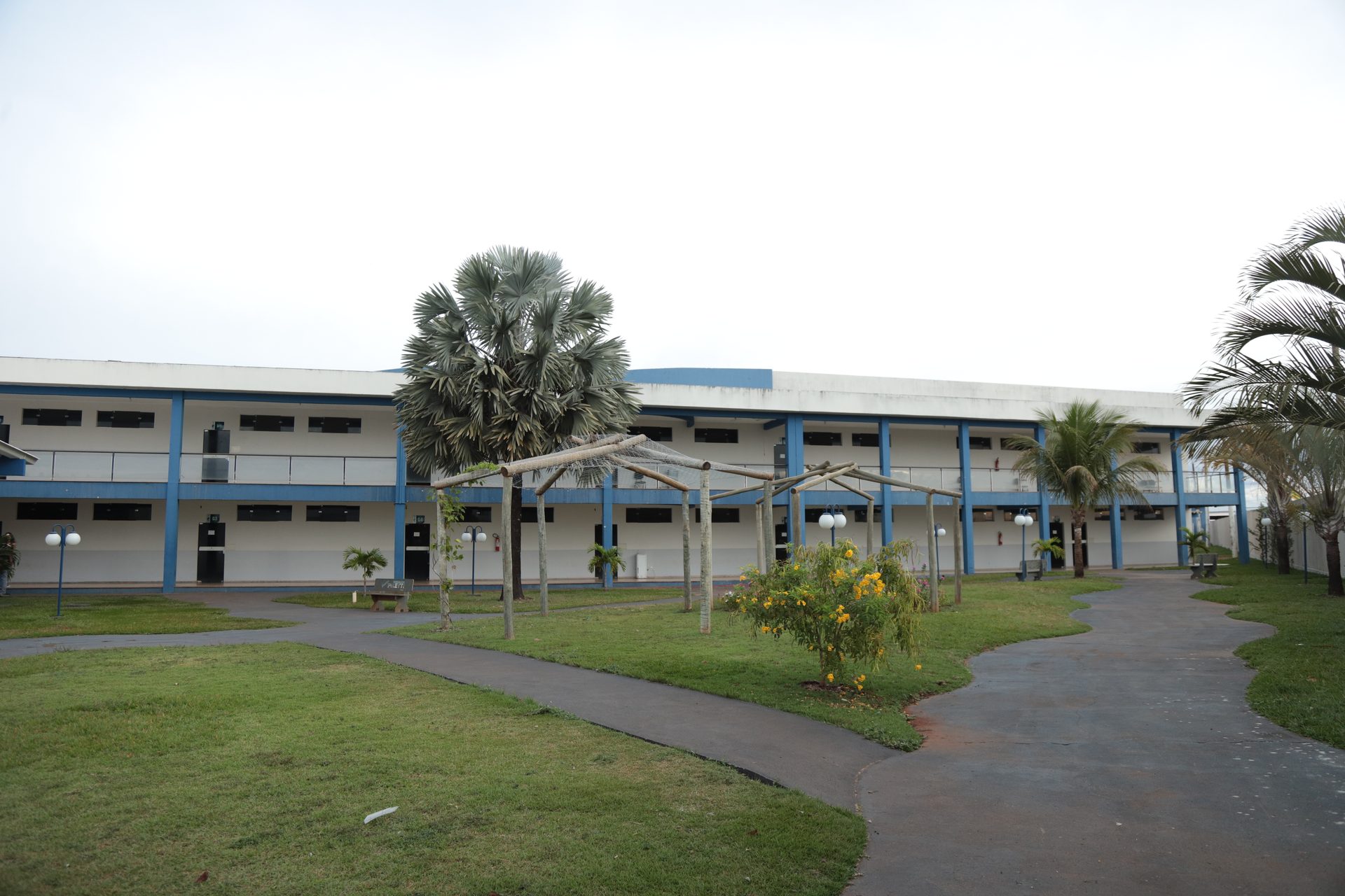 Vista externa e ampla de um prédio institucional de dois pavimentos, com fachada branca e detalhes em azul, corredores com colunas e varanda no andar superior.