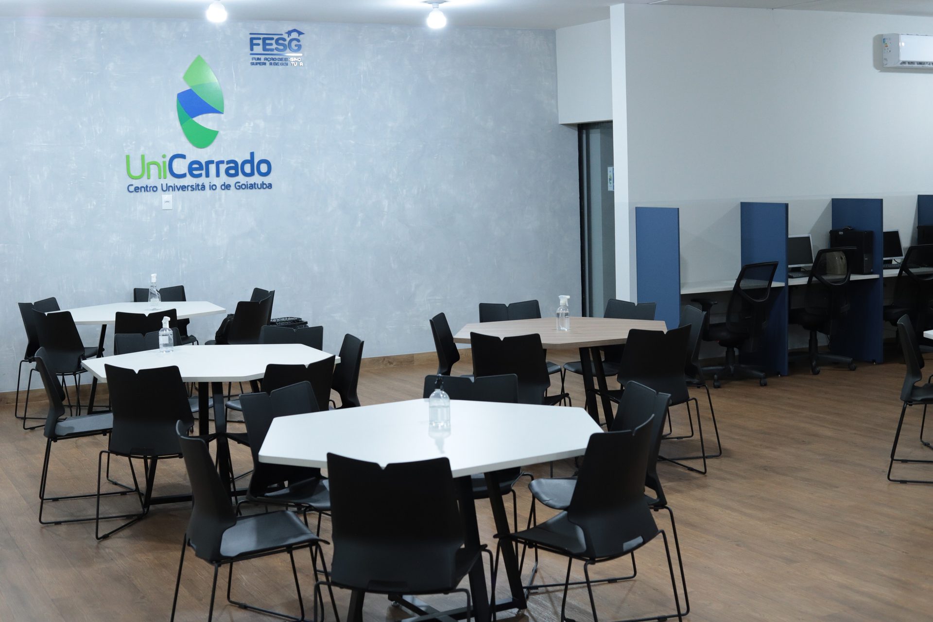 Ambiente amplo com piso de madeira, mesas hexagonais brancas com cadeiras pretas dispostas em grupos, cada mesa com uma garrafa de água.