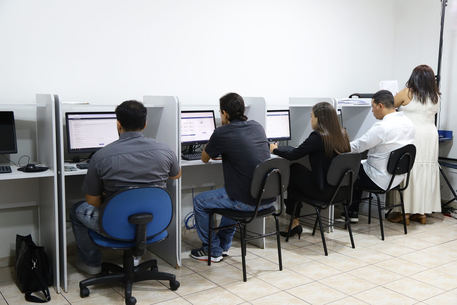 Ambiente com fileira de estações individuais tipo cabine, cada uma com computador de mesa (monitor, teclado e mouse), separadas por divisórias claras.