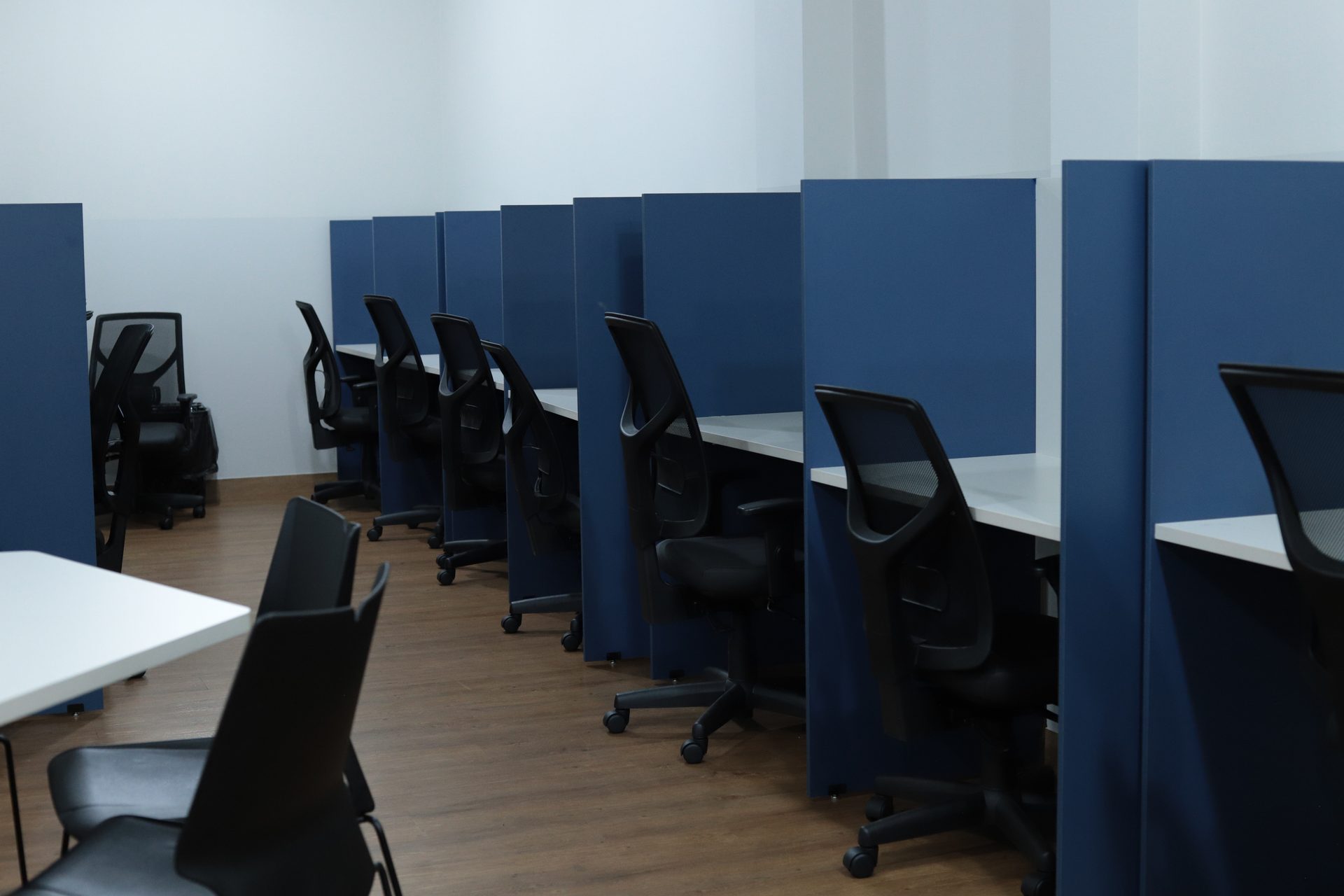 Ambiente vazio com fileira de baias de estudo individuais separadas por divisórias azuis, cada uma com mesa branca e cadeira giratória preta com rodízios.