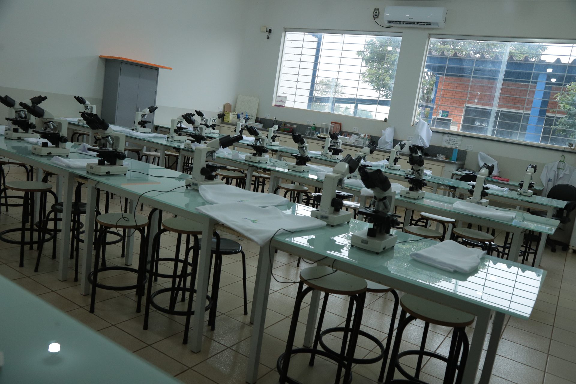 Laboratório vazio com fileiras de bancadas de superfície espelhada dispostas em diagonal, cada uma equipada com vários microscópios binoculares e jalecos brancos dobrados sobre as mesas.