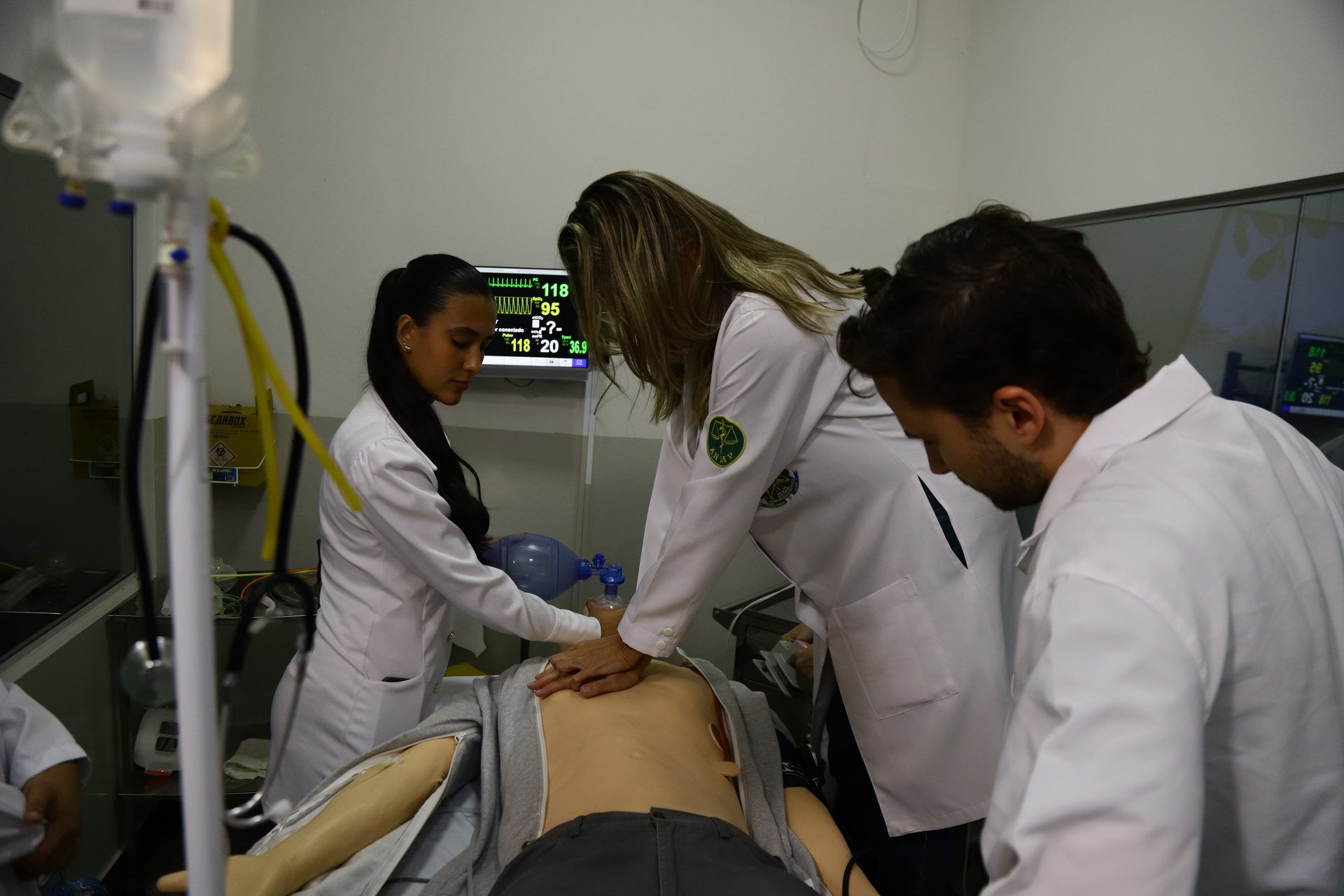 Três pessoas de jaleco branco realizam procedimento de simulação de emergência em um manequim de treinamento deitado em maca.