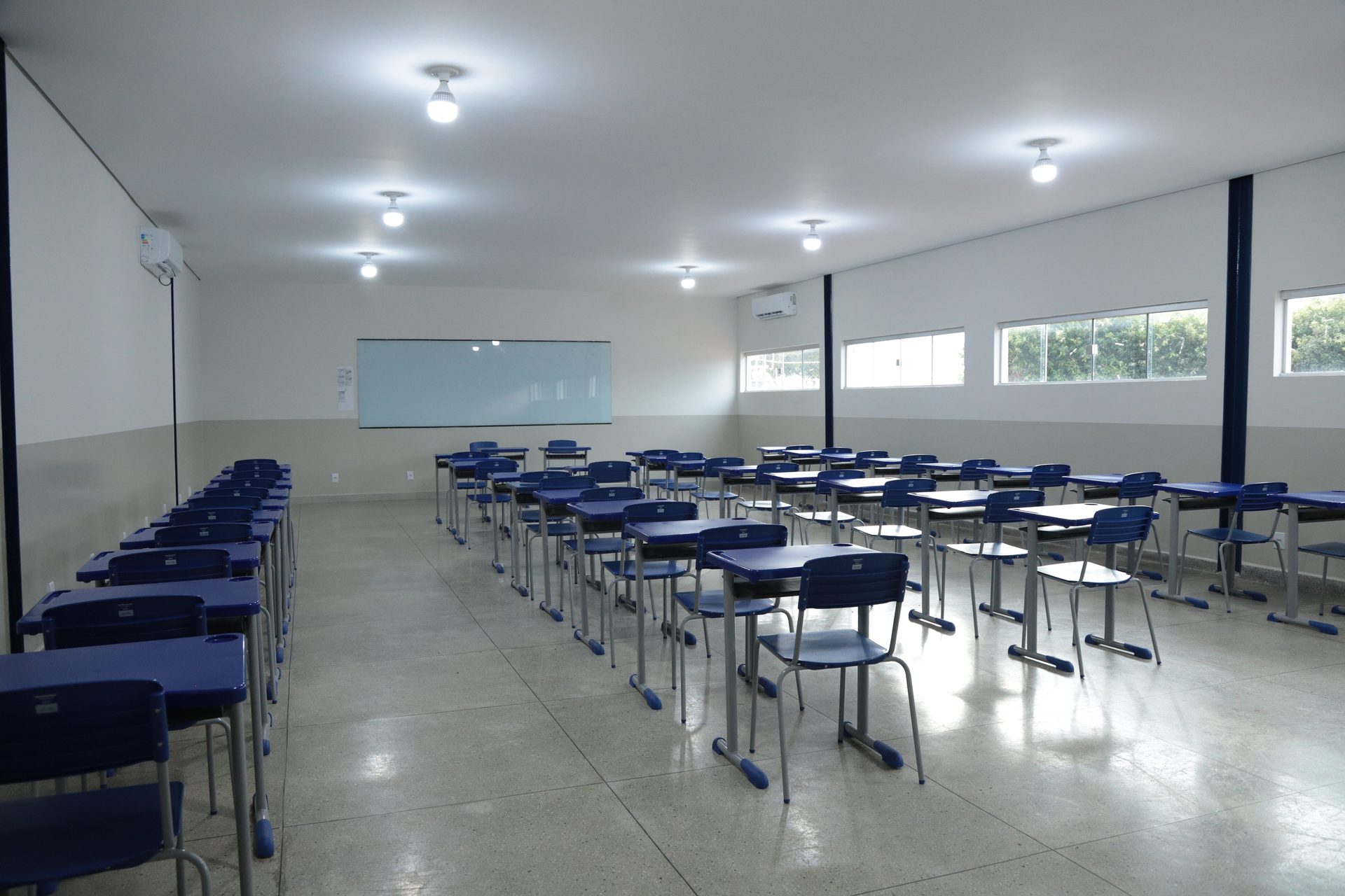 Sala de aula vazia com carteiras escolares azuis dispostas em fileiras sobre piso de porcelanato claro.
