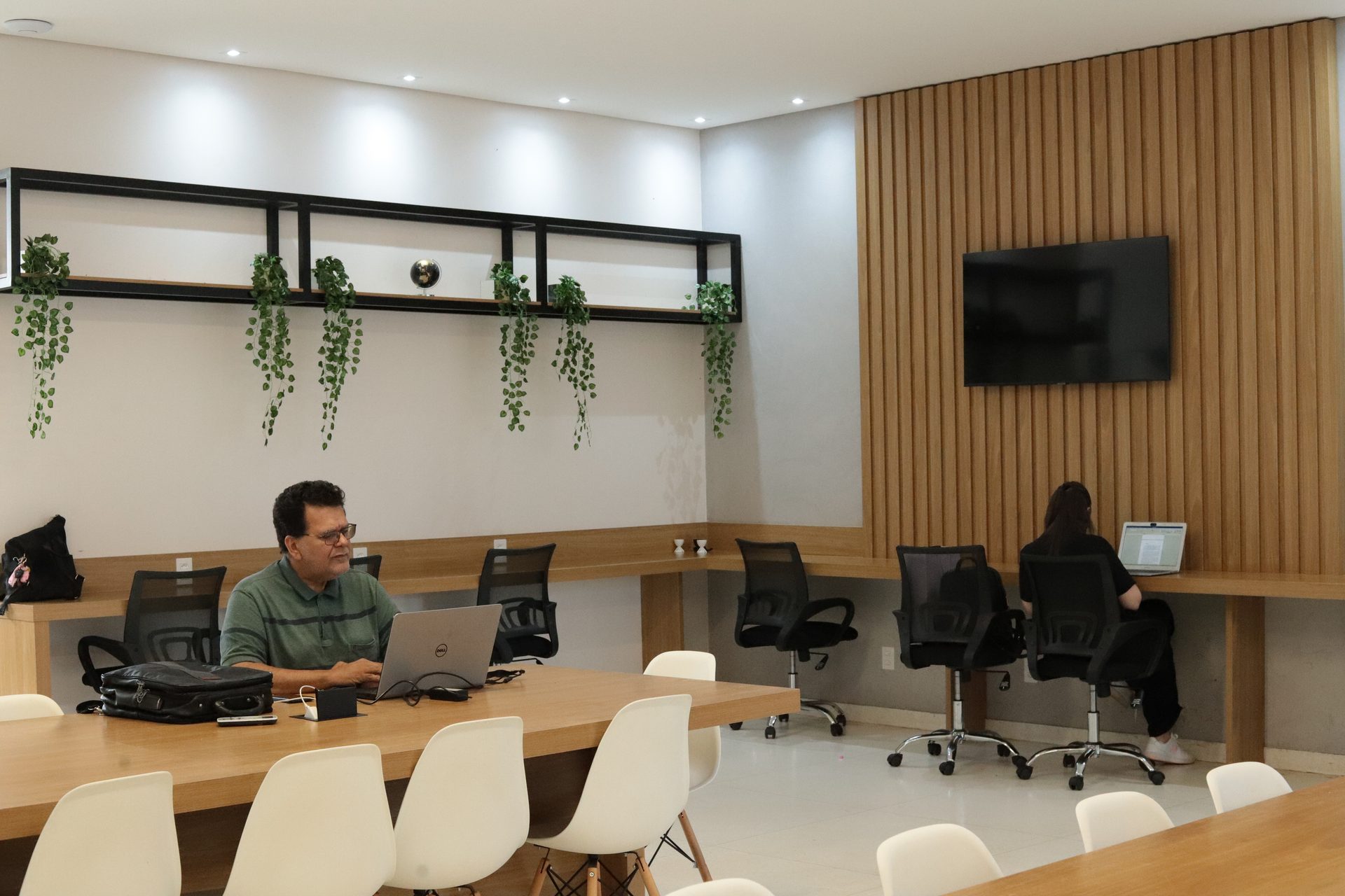 Ambiente de coworking com decoração moderna. Um homem de camisa verde está sentado à mesa de madeira em primeiro plano, usando um notebook, com uma bolsa ao lado.